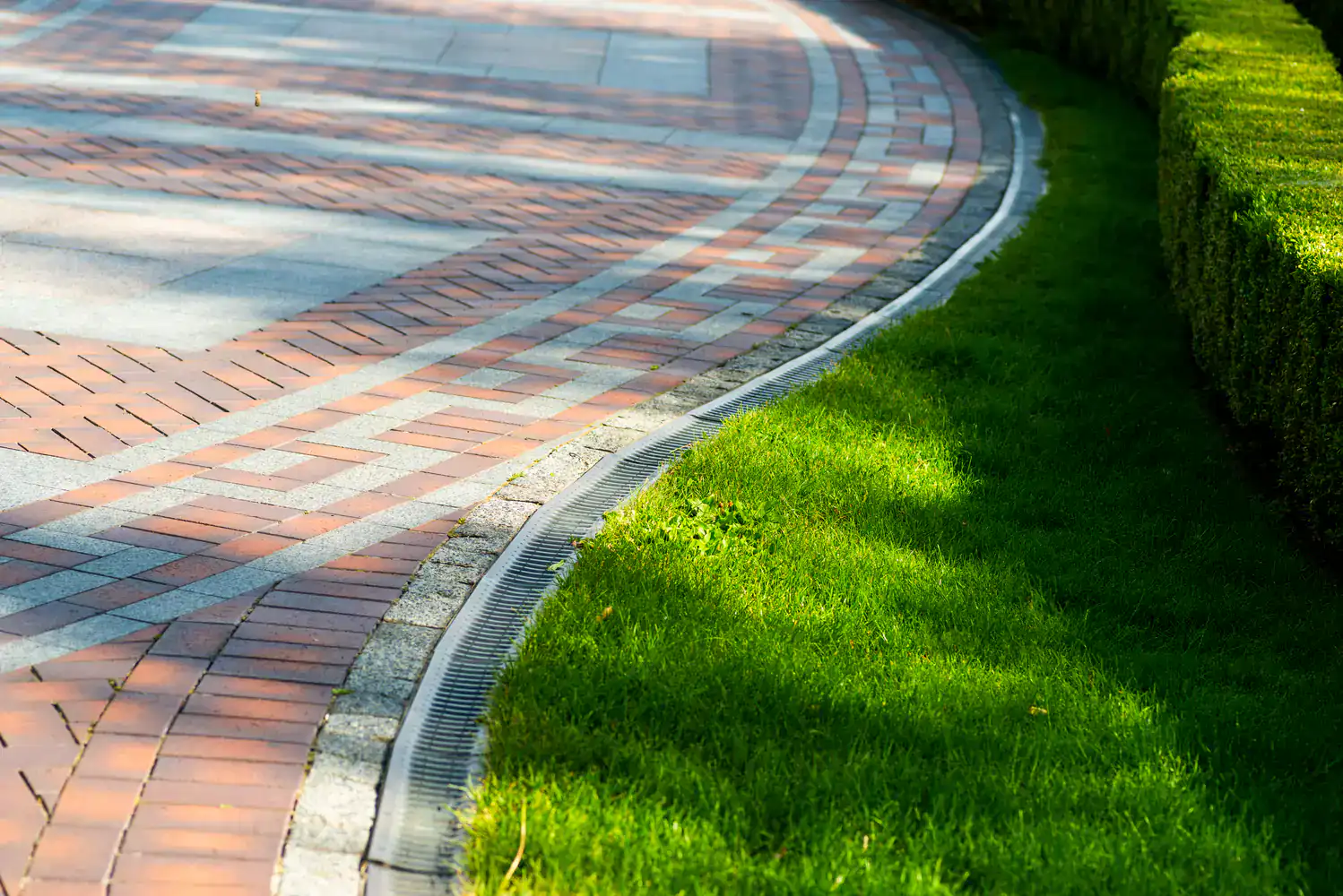 Curved brick pathway beside green grass and neatly trimmed hedges under bright sunlight.