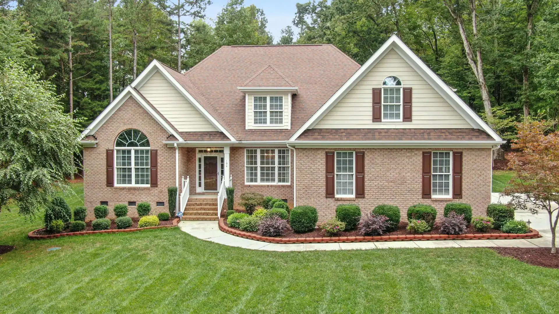 A large, two-story brick NY house with brown shutters stands behind a manicured lawn and landscaped shrubs.