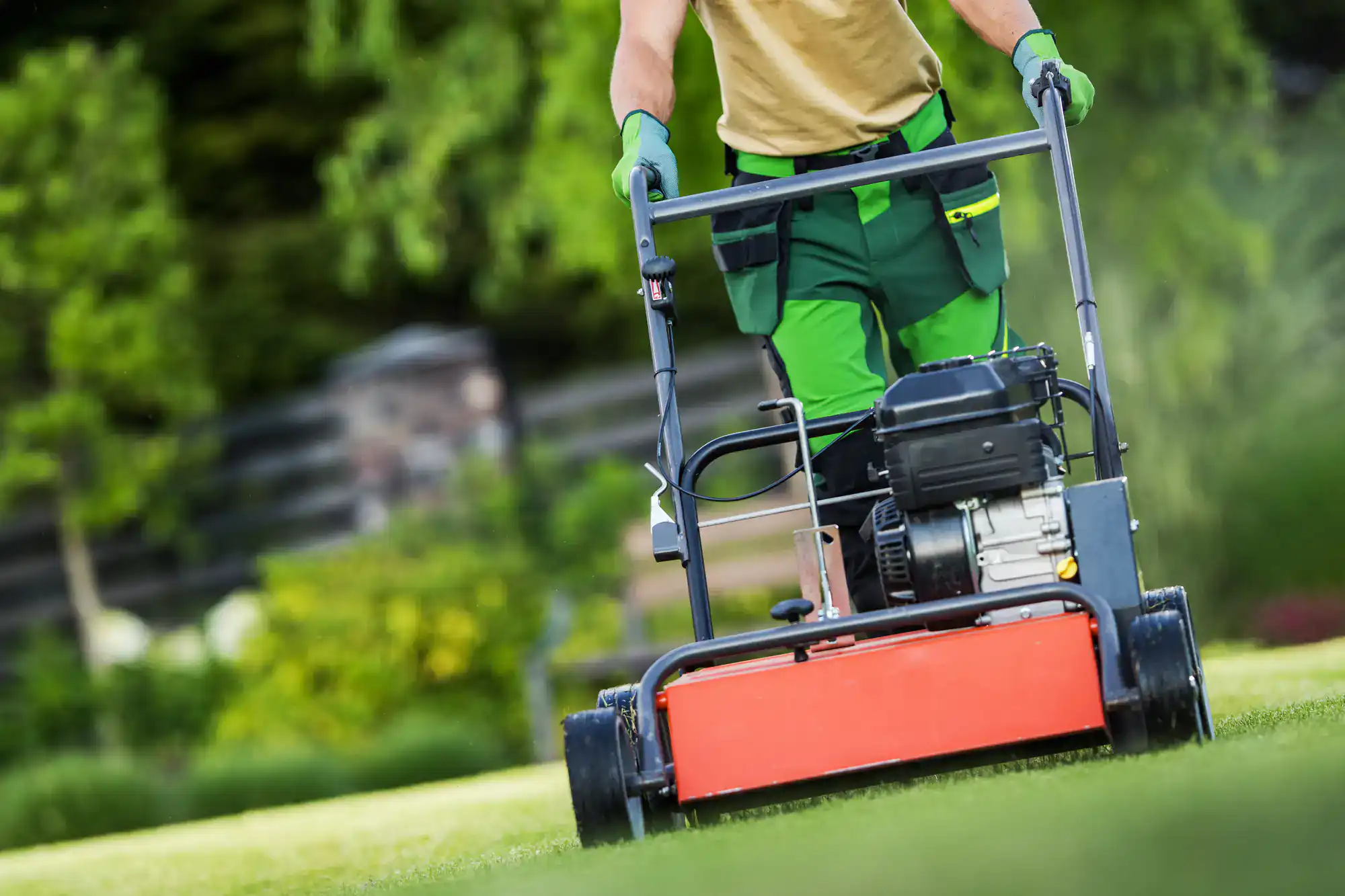 A Lawn Renovation Suffolk County expert mows a green lawn with a red mower, wearing green gloves.