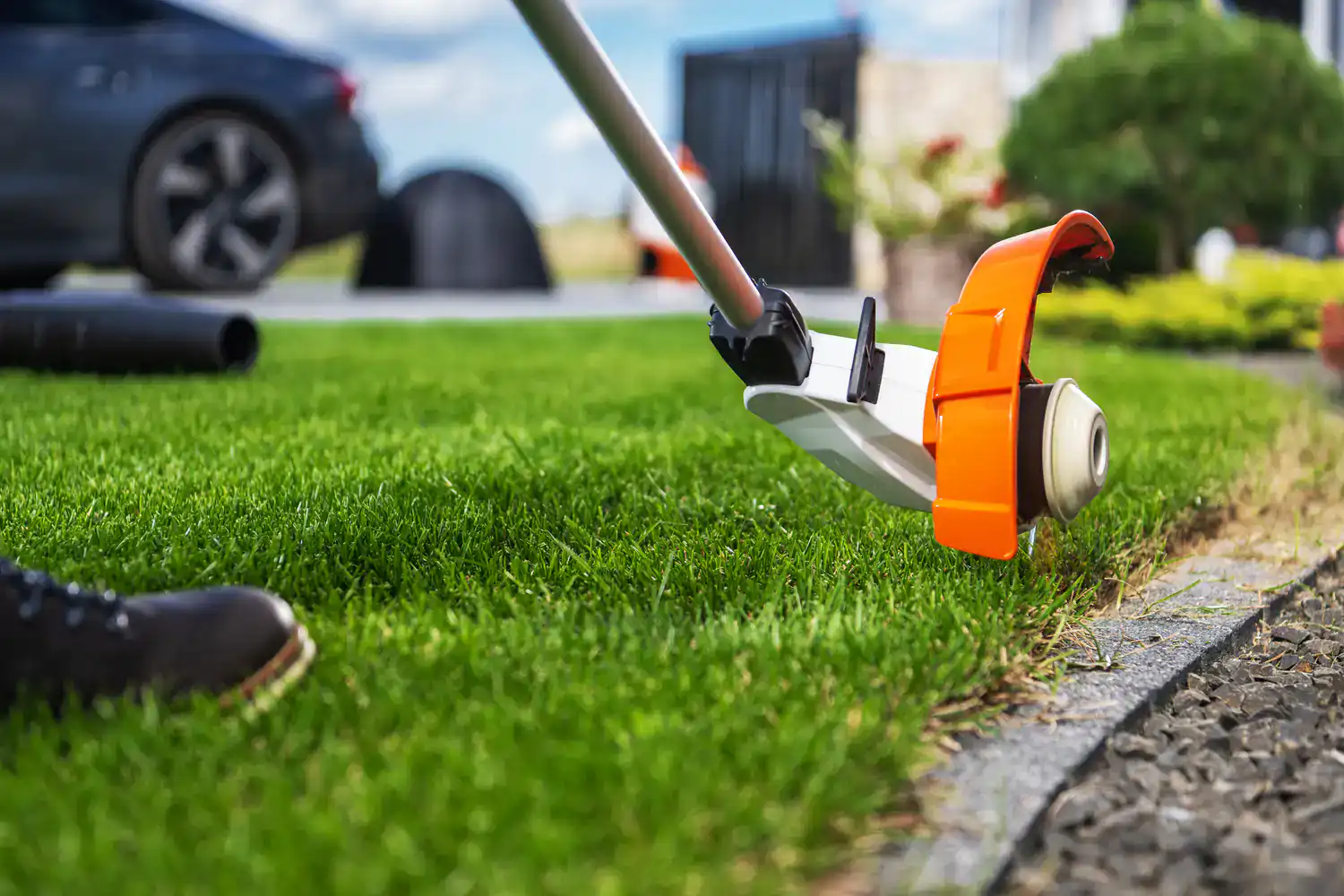 A person edges grass with an orange and white string trimmer along a stone walkway in their yard.