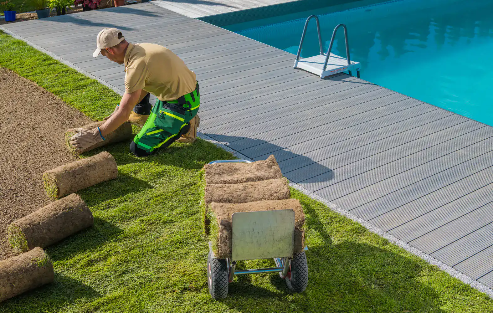 A worker installs sod grass beside a backyard pool, with a cart of rolled sod turf ready for landscaping.