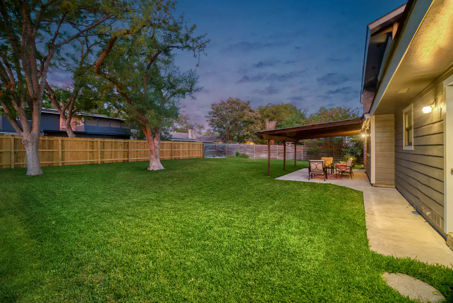 A backyard at dusk features green grass, trees, a wooden fence, and a covered patio with outdoor seating.