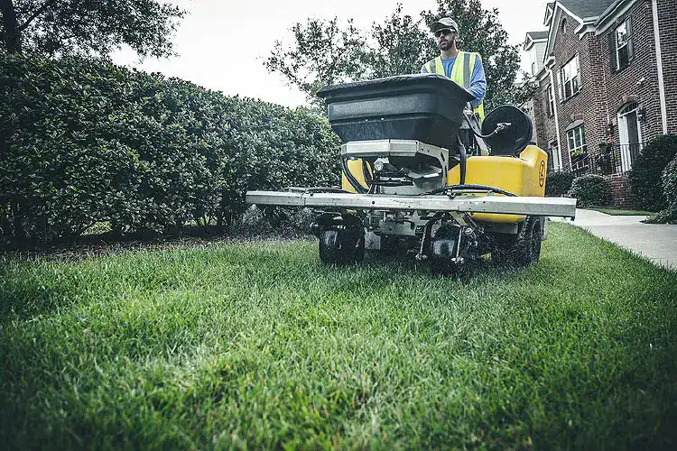 Worker driving a lawn care machine sprays grass near a hedge and brick house on a cloudy day.